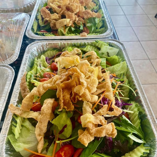 Two foil trays of Thai E-San salad with mixed greens, tomatoes, cucumbers, and crispy wonton strips.