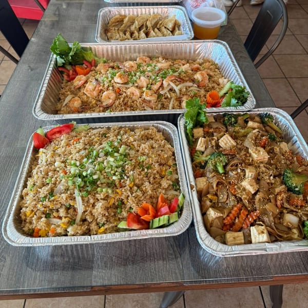 Three large catering trays of Thai food, including fried rice with shrimp, noodles with tofu, and dumplings.