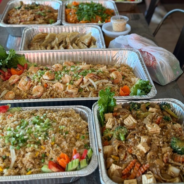 Multiple aluminum trays of Thai E-San dishes, including fried rice with shrimp, dumplings, and stir-fry.